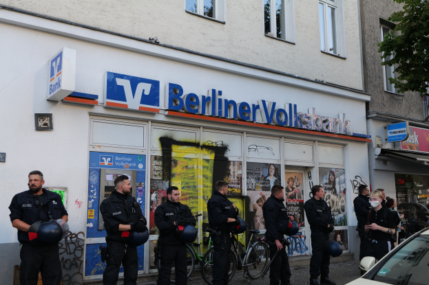 Eine Gruppe von Polizisten steht vor einer Berliner Volksbank, einige halten Helme, mit einem Auto rechts, einem Baum links und Gebäuden mit Fenstern und Texttafeln im Hintergrund.
