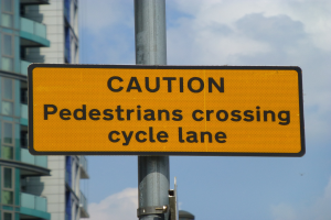 Gelbes Warnschild an einem Pfahl vor einem Gebäude, auf dem "Fußgänger überqueren Fahrradspur" steht.