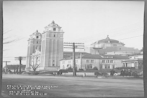 Ein Schwarz-Weiß-Foto einer römisch-katholischen Kathedrale und eines Bischofssitzes mit verschiedenen Gebäuden, Bäumen, Polen, Drähten, Menschen und einer Straße im Vordergrund und dem Himmel im Hintergrund, mit Text am unteren Rand des Bildes.