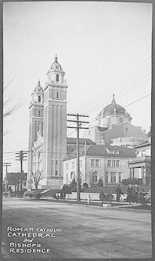 Ein Schwarz-Weiß-Foto einer römisch-katholischen Kathedrale und eines Bischofssitzes mit verschiedenen Gebäuden, Bäumen, Polen, Drähten, Menschen und einer Straße im Vordergrund und dem Himmel im Hintergrund, mit Text am unteren Rand des Bildes.