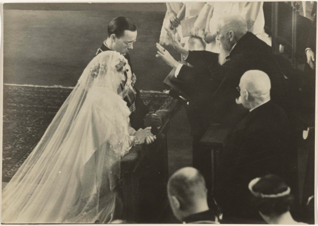 Ein Schwarz-Weiß-Foto einer Braut in einem weißen Kleid und Schleier mit einem Strauß Blumen in der Hand, die neben einem Bräutigam in einem Anzug und Krawatte steht, umgeben von einer Menschenmenge, einige klatschen.
