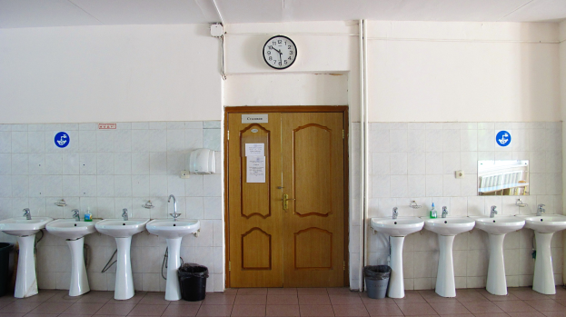 Reihe von Waschbecken mit Wasserhähnen, Mülltonnen auf dem Boden, einer Tür im Hintergrund, einer Uhr an der Wand und einem Spiegel auf der rechten Seite in einem Badezimmer.