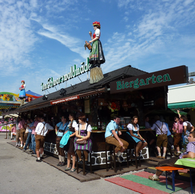 Eine Gruppe von Menschen vor einem Gebäude auf dem Oktoberfest in München, einige in bayrischer Tracht, andere auf Hockern sitzend, mit Tischen, Flaschen, einer Schiefertafel, Bäumen und bewölktem Himmel im Hintergrund.