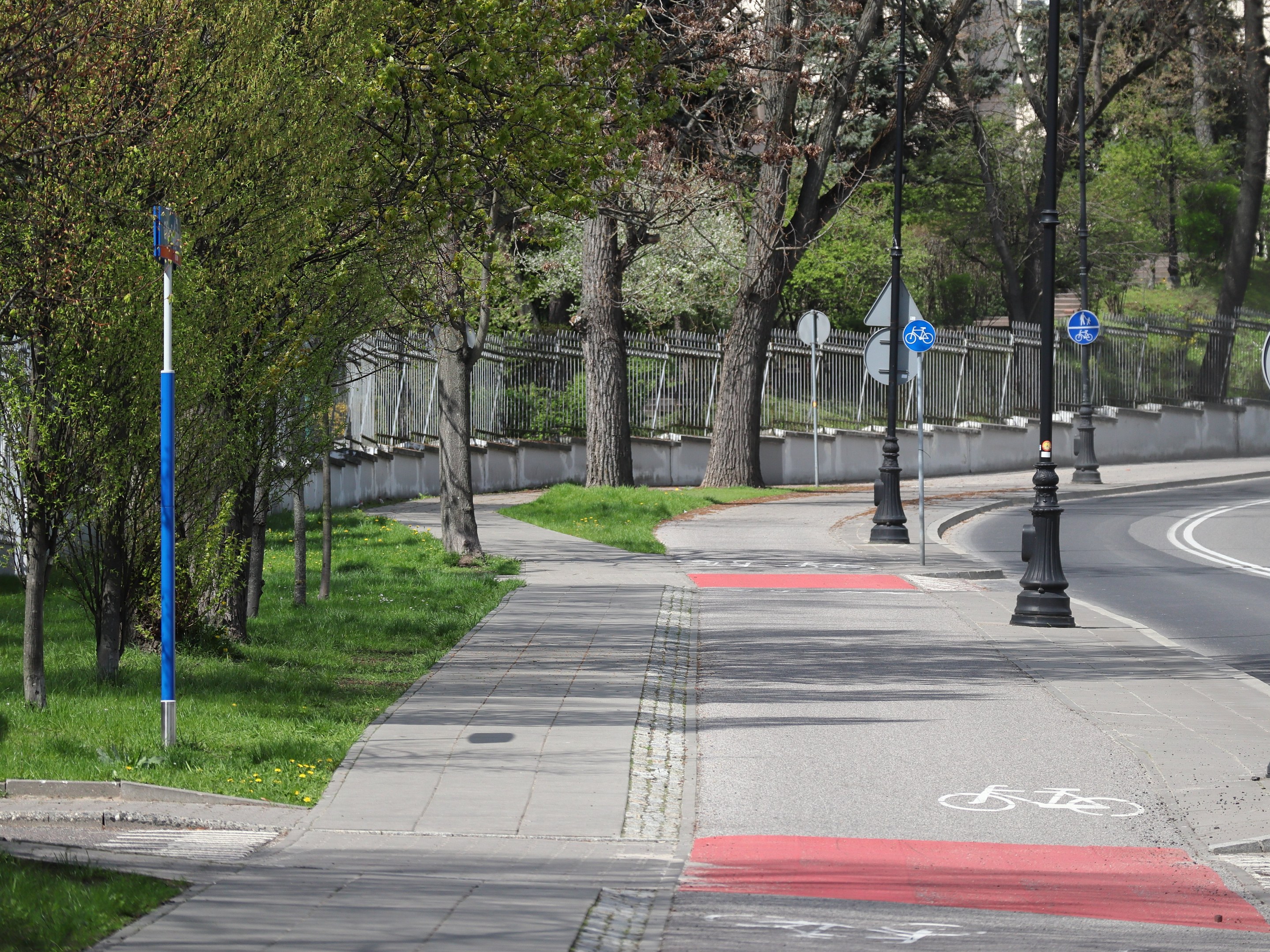 Stadtstraße mit einem Fahrradweg, Bäumen, Pfählen, Schildern, Gras, Pflanzen, einem Zaun, Gebäuden und einem klaren blauen Himmel im Hintergrund.