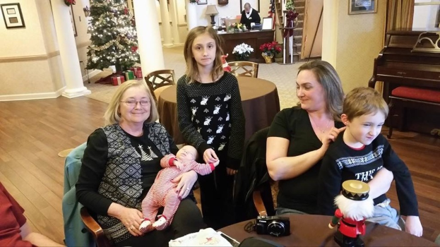 Ein Raum mit einer Wand, die ein Fotoframe, einen Weihnachtsbaum, eine Lampe und Pflanzen mit Blumen zeigt. Im Vordergrund gibt es Treppen, einige Menschen an Tischen mit einem Spielzeug und einer Kamera auf einem der Tische.