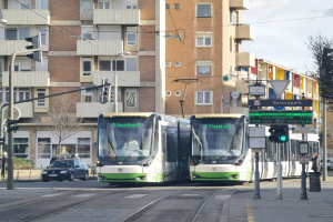 Zwei grüne und weiße Straßenbahnen auf einer Stadtstraße mit hohen Gebäuden, Fahrzeugen, Fußgängern, Ampeln, Pfählen, Schildern und Bäumen unter einem bewölkten Himmel.