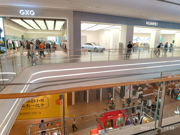 Ein Einkaufszentrumsinneres mit Menschen, die gehen, Glasgeländern, Bekleidungsauslagen, Informationsschildern, Säulen, Deckenleuchten und einem geparkten Auto im Hintergrund, mit einem GXG- und Huawei-Partnerschafts-Wasserzeichen.