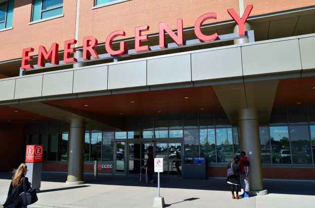 Eine Gruppe von Menschen vor einem Notfallgebäude mit Glaswänden, Säulen, einem Namensschild, Hinweistafeln auf Pfählen und einem Abfalleimer.