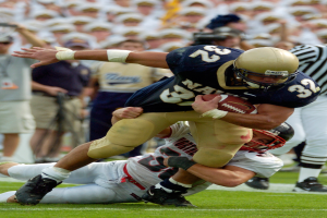Ein Fussballspieler in Sportkleidung und Helm wird von einem anderen Spieler auf dem Feld getackelt, mit anderen Spielern und Zuschauern im Hintergrund.
