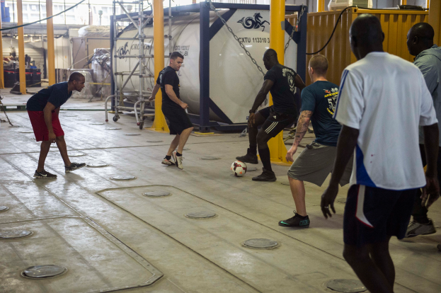 Eine Gruppe von Männern, die drinnen Fußball spielen, mit einem Ball auf dem Boden, Metallstangen, einem Container, einem Metallzaun, Stangen mit Ketten, einer Wand und einer Decke mit Lampen.