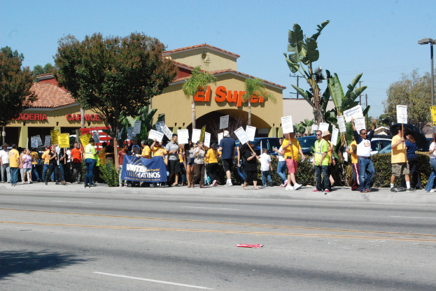 Eine Gruppe von Menschen mit Schildern und Bäumen, kleinen Büschen, einer Fahne, einem Gebäude mit Namensschildern und Autos im Hintergrund, möglicherweise während einer Wahl.
