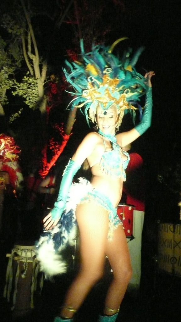 Eine Frau in einem eleganten Kostüm tanzt mit Trommeln und anderen Menschen im Hintergrund, mit einem Baum in der Nähe.