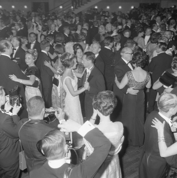 Schwarzes und weißes Foto von Menschen, die auf einem beleuchteten Tanzboden tanzen, einige halten Kameras, mit einer Treppe im Hintergrund.