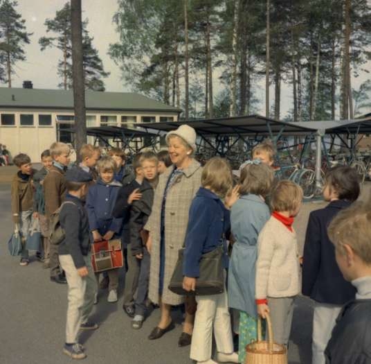 Eine Gruppe von Kindern steht vor einem Gebäude, einige halten Taschen und einer einen Korb, mit Fahrrädern, Schuppen, Bäumen und einem klaren blauen Himmel im Hintergrund, was an eine schwedische Schulszene der 1960er Jahre erinnert.