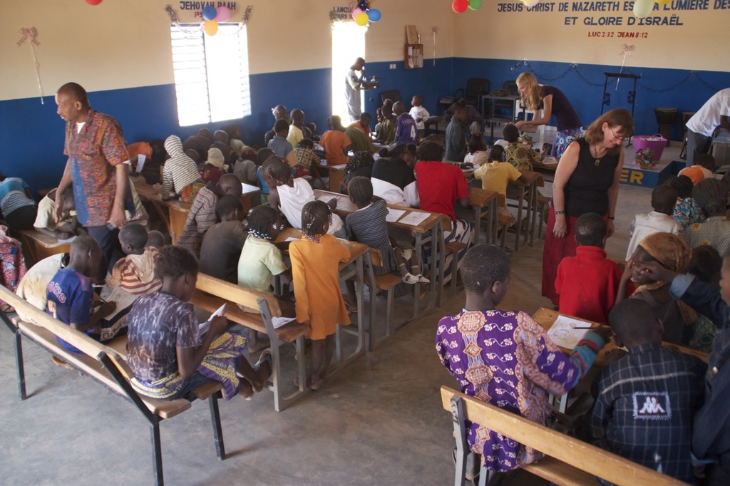 Kinder auf Bänken im Vordergrund mit begleitenden Erwachsenen, Stühle, eine Tür, ein Fenster und ein Tisch im Hintergrund, Ballons und Text oben.