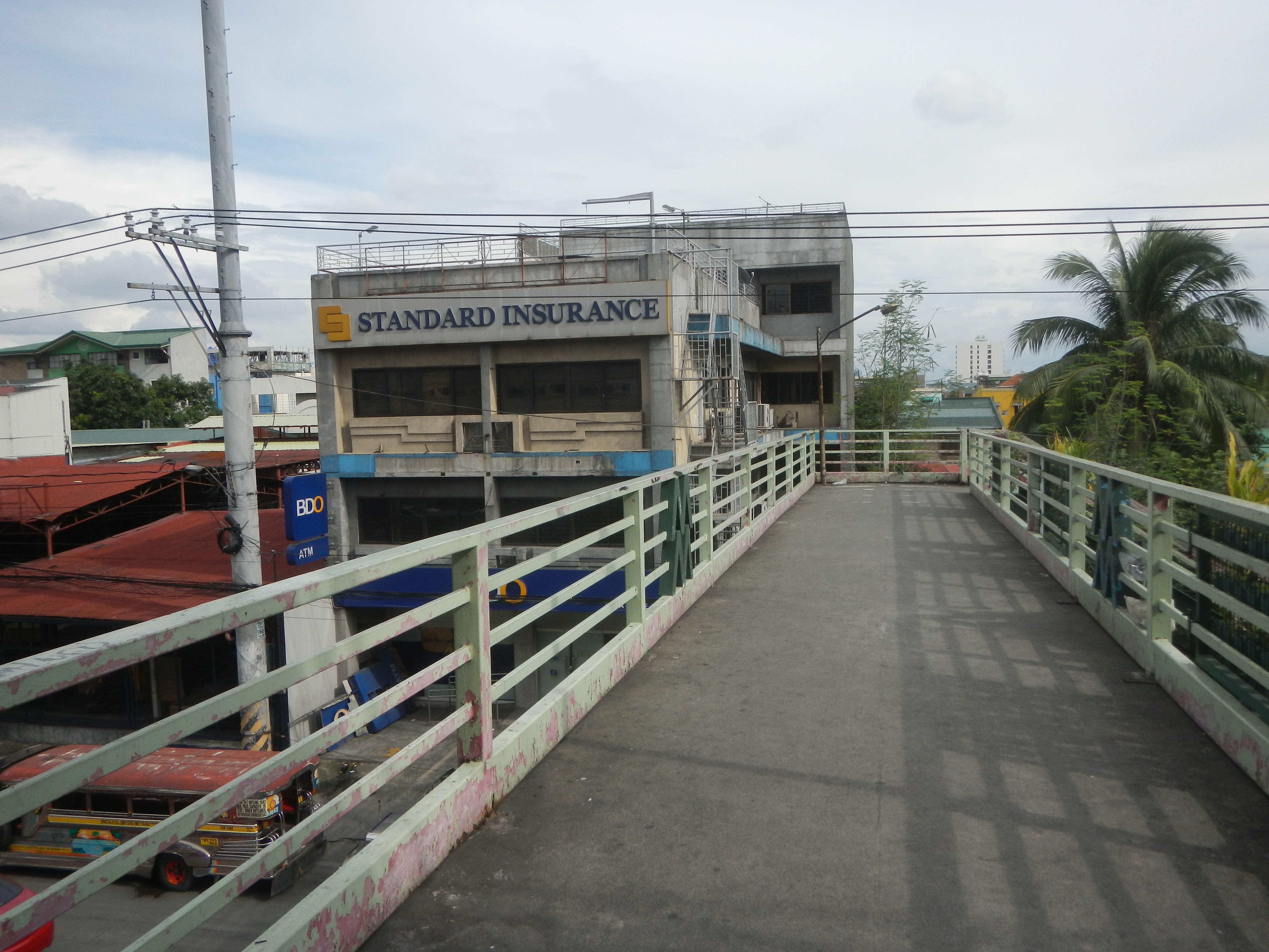 Eine Brücke mit Geländern, Fahrzeuge auf der Straße, Gebäude, Bäume, Strommasten mit Drähten und ein bewölkter Himmel im Hintergrund, mit einem Gebäude, auf dem eine Tafel mit der Aufschrift "Standard Versicherung" steht.