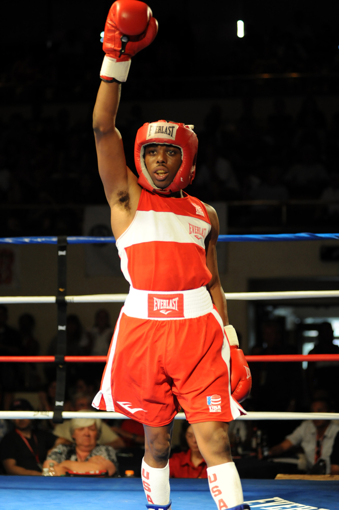 Ein Mann in Boxerkleidung, einschließlich Helm und Handschuhen, steht in einem Boxring mit Zuschauern im Hintergrund.