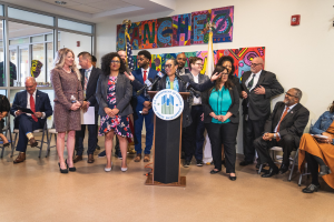Gruppe von Menschen vor einem Rednerpult mit Mikrofon und einer Tafel mit der Aufschrift "Sancho-Stadtrat tritt dem Stadtrat bei", mit sitzenden und stehenden Zuschauern, Flaggen, Feuerlöscher und Deckenleuchten im Hintergrund.