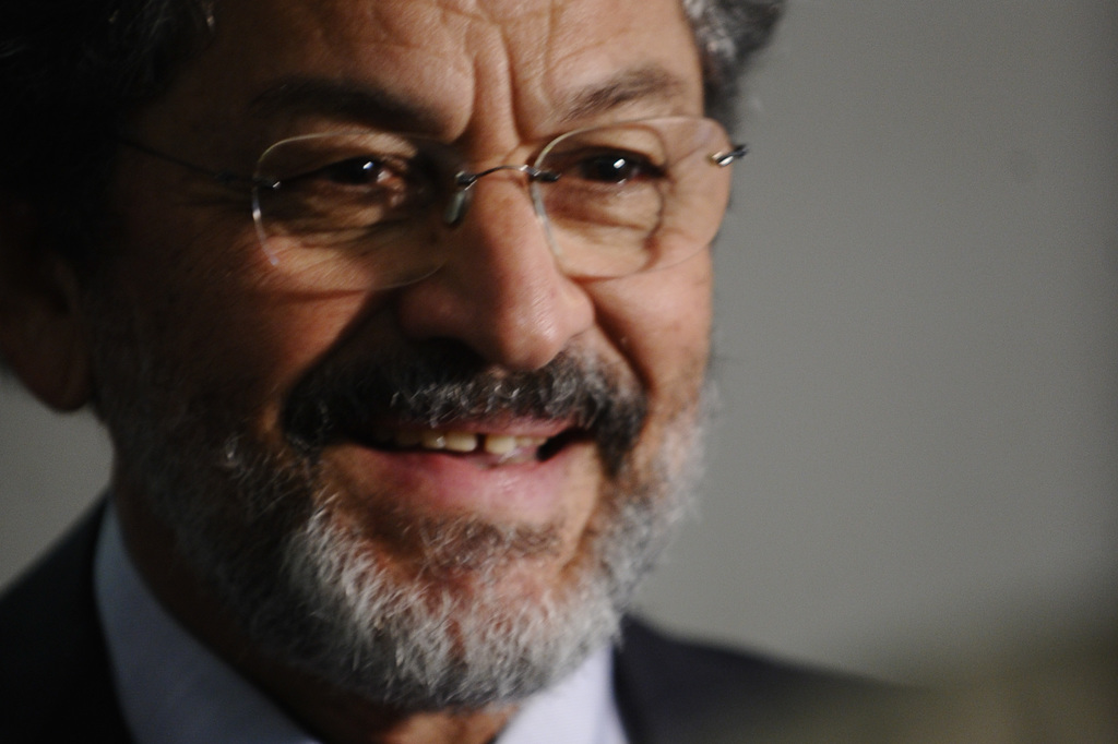 Ein alter Mann in einem schwarzen Anzug und weißem Hemd, der eine Brille trägt, lächelt.