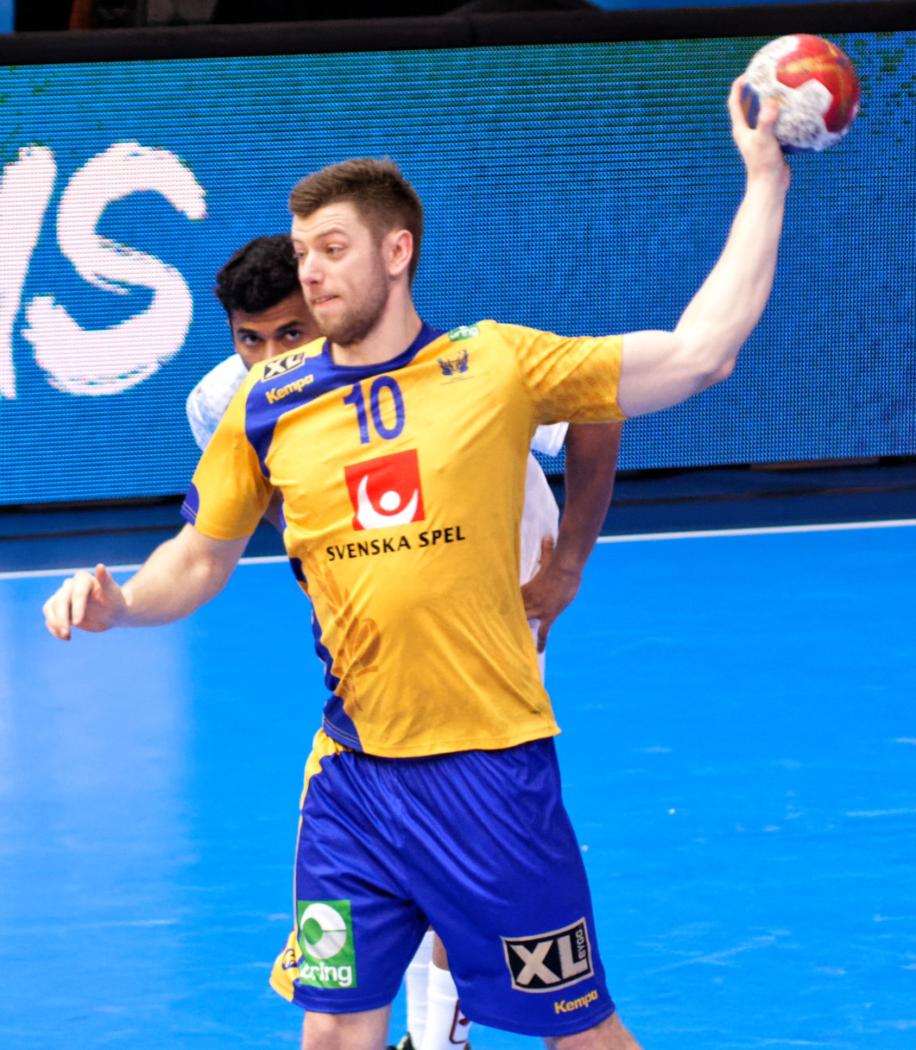 Ein Mann in einem gelben Hemd und blauen Shorts spielt Handball auf einem Platz, hält einen Ball und hat im Hintergrund einen Bildschirm mit der Aufschrift "Svenský Spel Handball World Cup 2019".