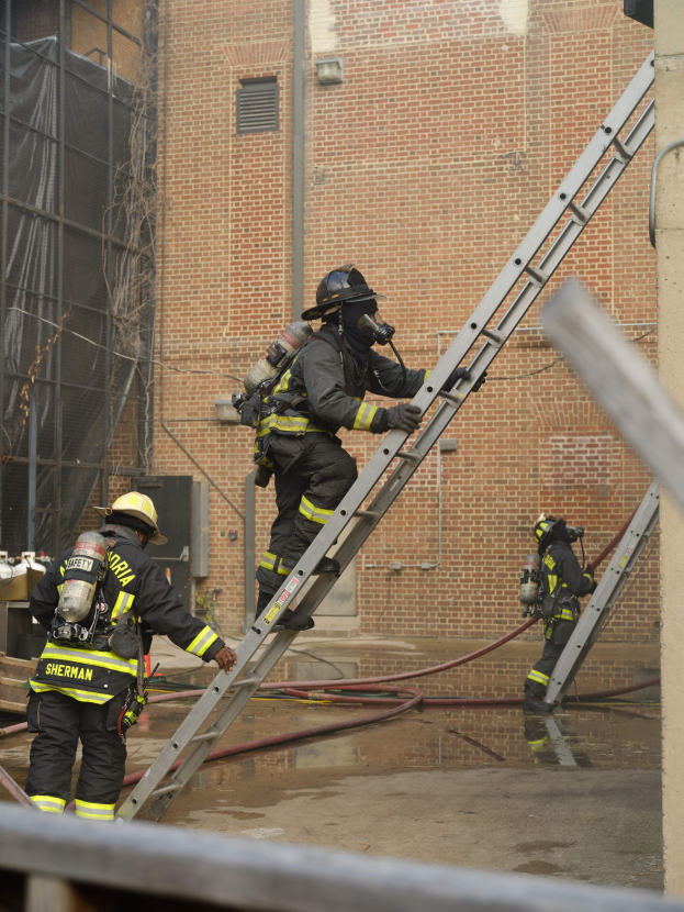 Feuerwehrleute mit Helmen und Luftzylindern klettern an einer Leiter vor einem Backsteingebäude mit Rohren auf dem Boden und einer Metallstange am Boden, mit einem weiteren Gebäude mit Fenstern und einem Netz im Hintergrund.