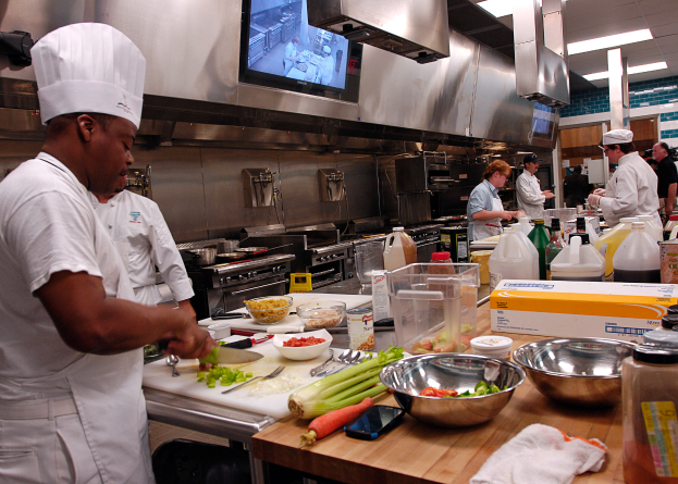 Eine Gruppe von Köchen in einer Küche beim Zubereiten von Speisen, mit einem Koch im Vordergrund in weißer Kleidung und Mütze, der ein Messer hält und von verschiedenen Utensilien, Schüsseln, Gemüse, einem Handy, Flaschen und anderen Gegenständen umgeben ist, während im Hintergrund ein Fernseher zu sehen ist.
