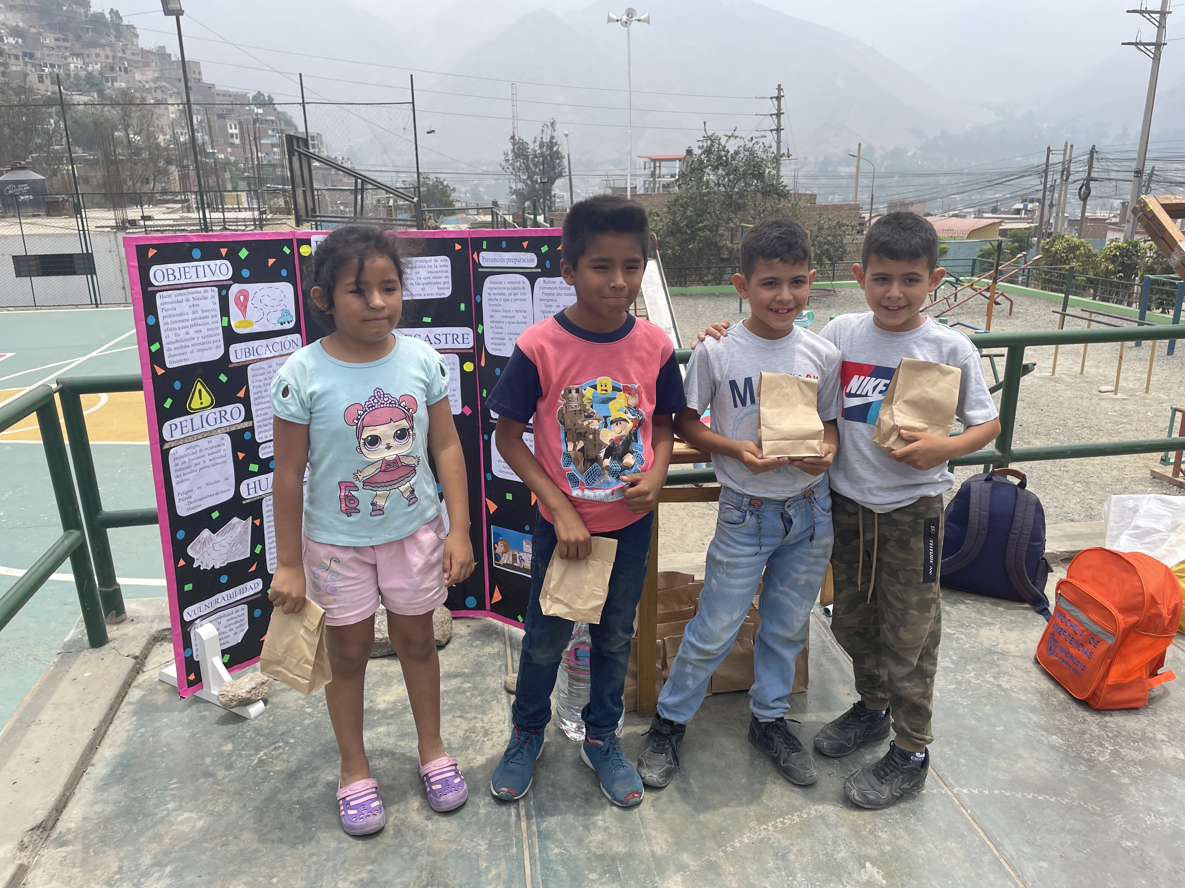 Eine Gruppe von Kindern steht auf einem Basketballfeld, jedes Kind hält eine Papiertüte in der Hand, rechts von ihnen liegen Tüten und Gegenstände auf dem Boden und links eine Tafel mit Text, im Hintergrund gibt es Zäune, Pfosten, Straßenlaternen, Strommasten, Bäume, Gebäude, Hügel und Himmel.
