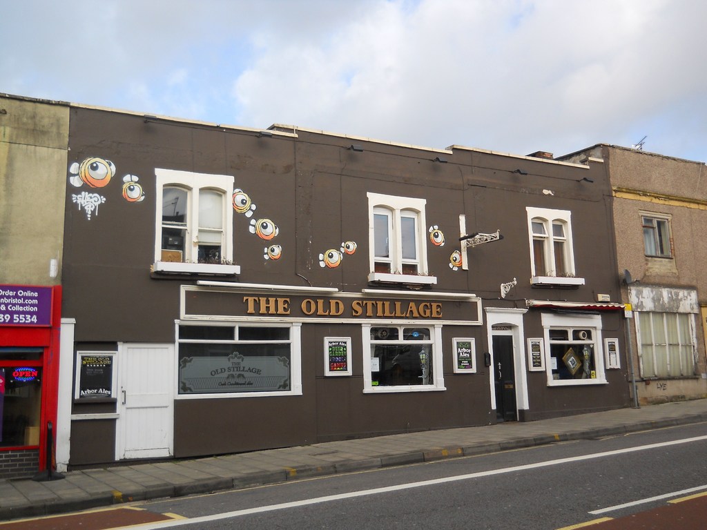 Eine Straßenszene außerhalb der Stadt mit einer Straße, einem Gehweg und Gebäuden mit sichtbaren Türen und Fenstern, einschließlich einer roten Wand und einem violetten Schild mit Text, unter einem klaren Himmel.
