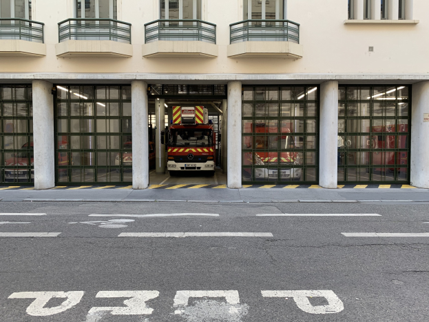 Feuerwache mit Feuerwehrauto davor, mit Fenstern, Geländern, Säulen, Glastüren und einer Straße unten.