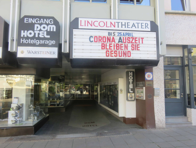Das Lincoln Theater in Berlin, Deutschland, ein Gebäude mit Glasfenstern und -türen und einer Tafel mit Text darauf, das verschiedene Objekte zeigt und den Eindruck einer pulsierenden Stadtlandschaft vermittelt.