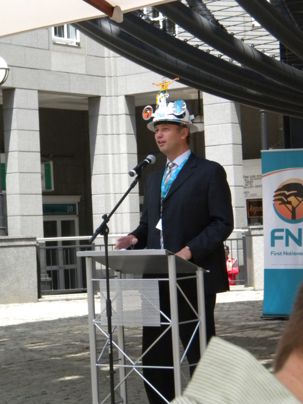 Ein Mann im Anzug und Hut spricht in ein Mikrofon an einem Podium, mit einer Person rechts unten, einem Banner mit Text rechts, einem Gebäude und einem Geländer im Hintergrund.