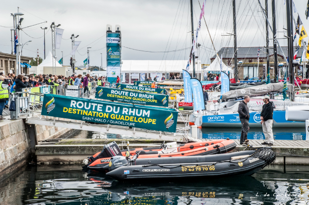Eine belebte Szene mit zahlreichen Booten, Bannern, Pfählen und Drähten, einer großen Menge hinter einer Absperrung auf der linken Seite und zwei Personen auf einer Brücke über dem Wasser, mit Abteilen um die Boote herum.