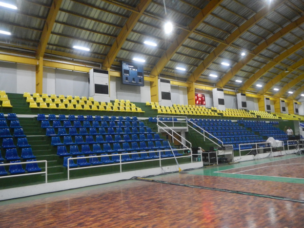 Große Sporthalle mit Holzboden, blauen und gelben Sitzplätzen, Geländern, Treppen mit Geländern, Wandbildschirmen, Deckenleuchten und einigen anwesenden Personen.