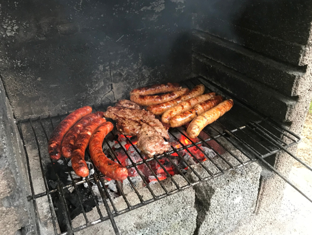 Grill mit Würstchen und Fleisch darauf, umgeben von brennender Kohle und Flammen, draußen auf Gras