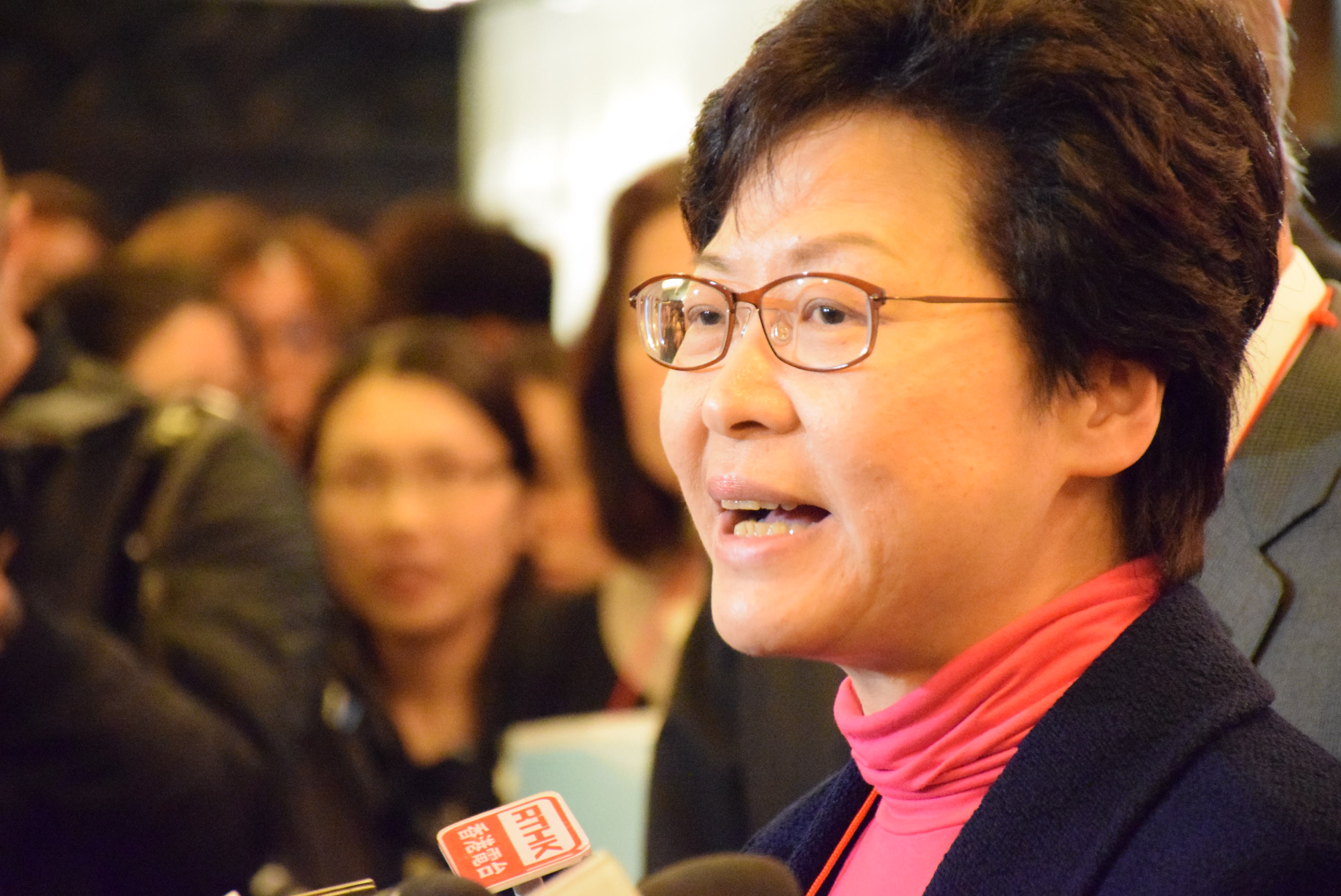 Eine Frau mit Brille und pinker Schärpe spricht mit Reportern, einige halten Mikrofone, mit einem unscharfen Hintergrund.