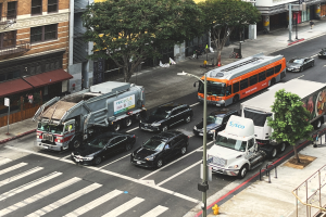Eine belebte Stadtstraße mit verschiedenen Fahrzeugen, Fußgängern, Gebäuden, Bäumen und Straßenmöblierung, darunter Geländer, Treppen, Laternenmasten und Schilder.