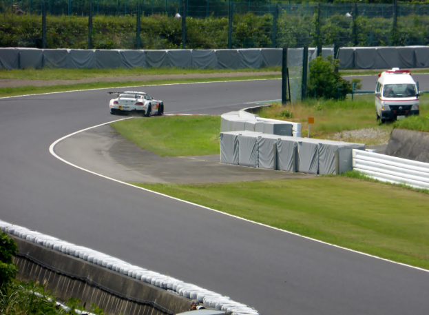 Ein Rennwagen fährt auf einer Rennstrecke mit einem Polizeiwagen im Hintergrund, umgeben von Gras, Pflanzen, Bäumen und Absperrungen, mit Text am unteren Bildrand.