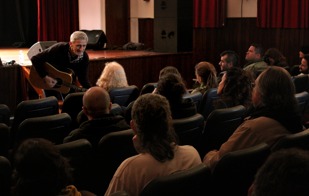 Ein älterer Mann in einem schwarzen Pullover spielt Gitarre auf einer Bühne mit Lautsprechern und einem Vorhang im Hintergrund, während viele Menschen auf Stühlen sitzen und ihm gegenüber.