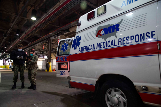 Ein Rettungswagen der American Medical Response (AMR) steht in einem Gebäude, vor ihm stehen zwei maskierte Personen; links ist ein Fahrzeug zu sehen, im Hintergrund sind weitere Personen, Lichter und Rohre.