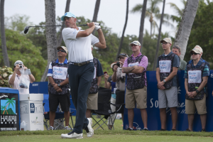 Ein Mann in einem weißen T-Shirt, schwarzen Hosen und einer blauen Mütze schwingt einen Golfschläger auf einem saftig grünen Feld, mit Zuschauern und Kameras dahinter unter einem klaren blauen Himmel.