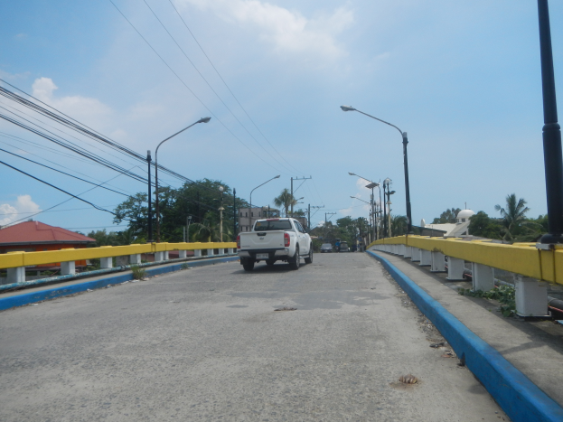 Ein Truck fährt über eine Brücke mit gelben Geländern, Laternenmasten, Strommasten mit Drähten, Bäumen, Gebäuden und einem bewölkten Himmel im Hintergrund.