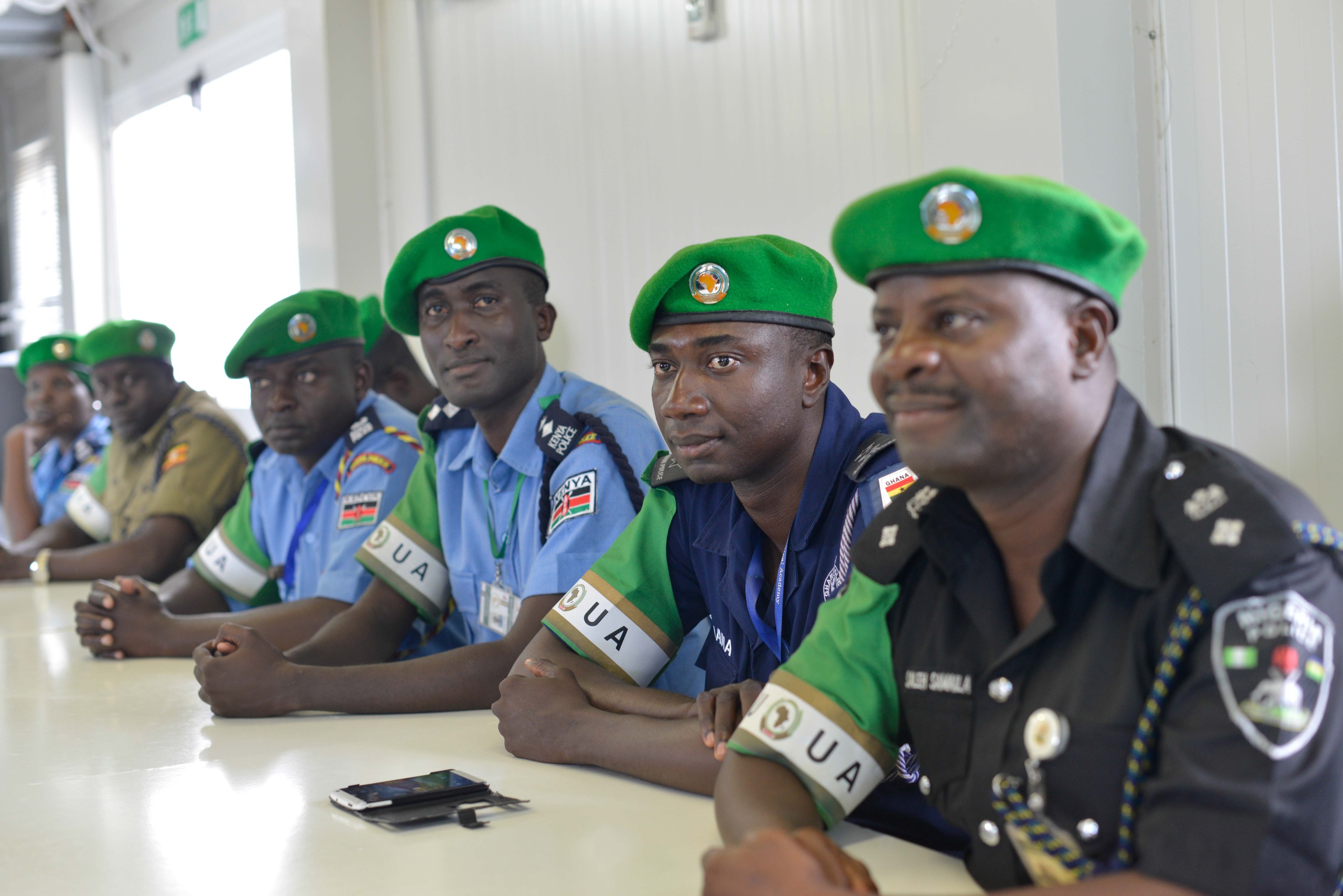 Polizisten sitzen an einem Tisch während einer Schulung, mit Mobiltelefonen und anderen Gegenständen auf dem Tisch, in einem Raum mit Fenstern und Deckenbeleuchtung.