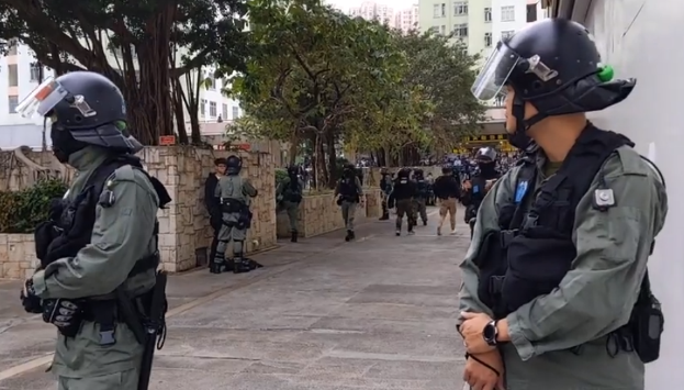 Eine Gruppe von Polizisten in Helmen und kugelsicheren Westen steht auf einer Hongkonger Straße, mit Bäumen, Gebäuden und einem klaren Himmel im Hintergrund.