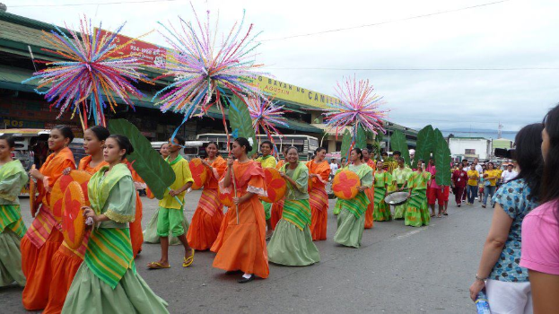 Eine Gruppe von Menschen in bunten Kostümen spaziert über eine Straße, während eines Karnevalsumzugs, mit einer Frau auf der rechten Seite, die eine Flasche hölt, und Fahrzeugen, Gebäuden, Bäumen, Polen, Dröhnen und Himmel im Hintergrund.