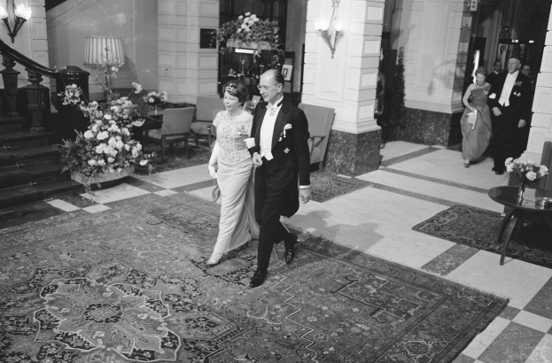 Schwarz-weiß-Foto einer Braut und eines Bräutigams, die durch die Lobby eines Hotels gehen, umgeben von Gästen, einem Tisch mit einer Blumenvase, Stühlen, Topfpflanzen, Lampen, einer Treppe mit Geländer und einer Wand.