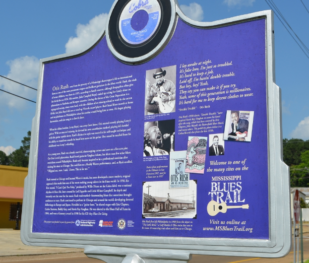 Ein Informationsschild, Gebäude, ein Schuppen, Bäume, Stromkabel und ein bewölkter Himmel.