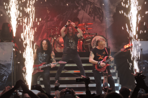 Eine Gruppe von Menschen, die auf der Bühne eines Konzerts spielen, darunter einige mit Gitarren, einer mit Mikrofon und einer, die im Hintergrund Schlagzeug spielt, während unten eine Menge zuschaut.