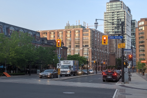 Eine stark befahrene Stadtstraße mit einem Lastwagen, einem Auto und einem SUV, gesäumt von Laternen, Ampeln, Anzeigetafeln, Bäumen und Gebäuden unter einem Himmel.