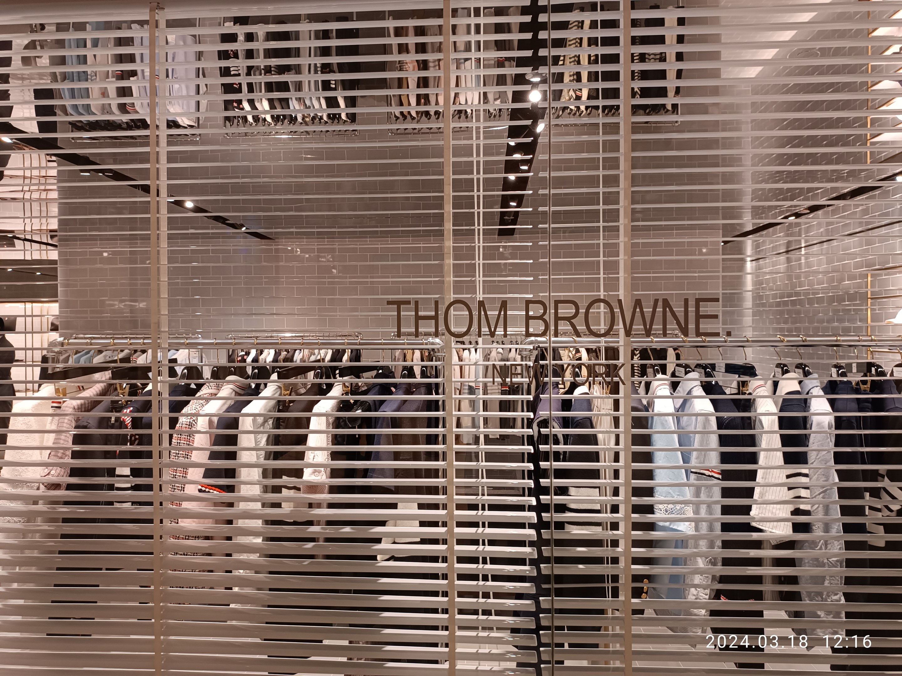Bekleidungsgeschäftsinnere mit Kleiderstangen, "Thom Browne"-Schilder, Deckenleuchten und Wasserzeichen.