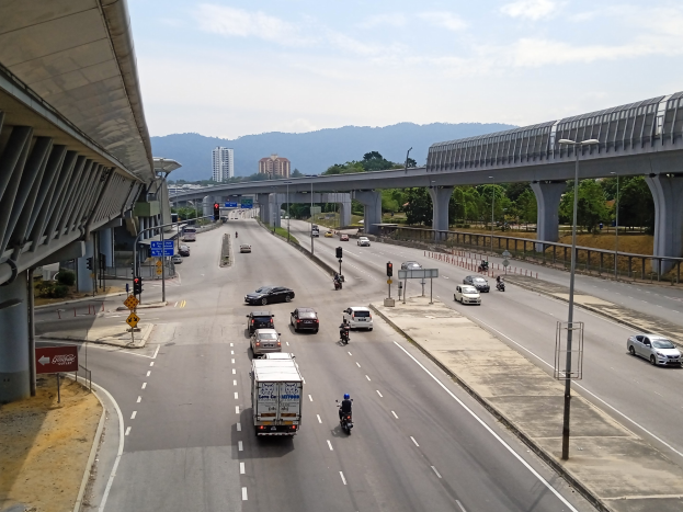Eine vielbefahrene Autobahn mit mehreren Fahrzeugen in der Nähe einer Brücke, mit Straßenlaternen, Ampeln, Schildern, Bäumen, Gebäuden, Hügeln und einem bewölkten Himmel.